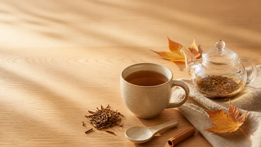 A warm mug of hojicha tea with roasted green tea leaves, autumn leaves, and a glass teapot on a sunlit wooden table